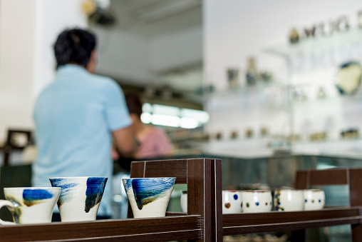 A saleswoman points at a plate being sold in a display of homemade pottery at a local small business owners shop.  Various cups and mugs can be seen in the back ground on glass display shelves.A saleswoman points at a plate being sold in a display of homemade pottery at a local small business owners shop.  Various cups and mugs can be seen in the back ground on glass display shelves.