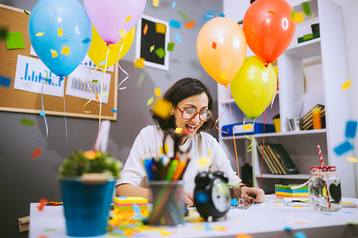 Business Might Be Fun If You Out To Celebrate Your Birthday At The Office. This Brunette Woman Looks Pleased And Happy While Sitting At The Multicolored Desk Full Of Balloons.