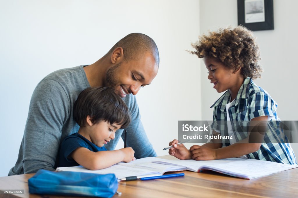 Father helping his sons with homework Young black father helping his boys with homework. Multiethnic brothers studying with their dad at home. Two cute children doing homeworks with the help of their loving parent. Father Stock Photo