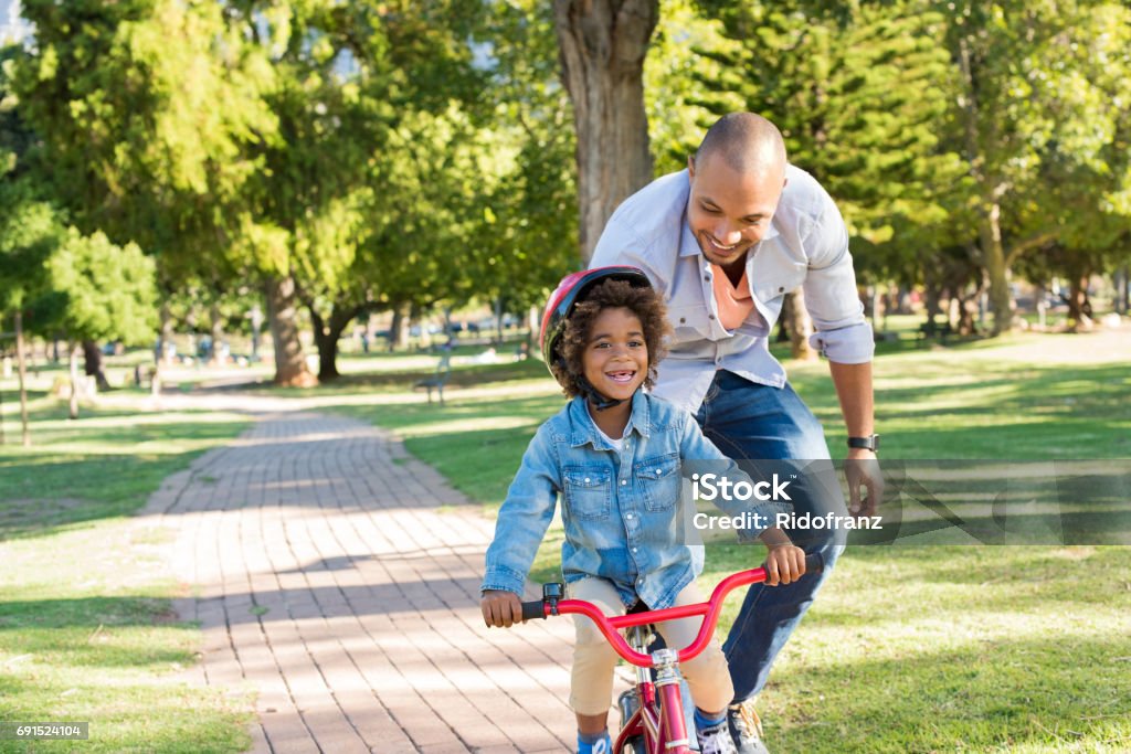 Father teaching son cycling Lovely father teaching son riding bike at park. Happy father helping excited son to ride a bicycle in a summer day. Young smiling black boy wearing bike helmet while learning to ride cycle with his dad. Cycling Stock Photo