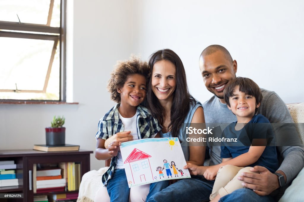 Multiethnic family on sofa Proud parents showing family painting of son sitting on sofa at home. Smiling mother and father with children"u2019s drawing of a new home. Black little boy with his family at home showing a painting of a happy multiethnic family. Family Stock Photo