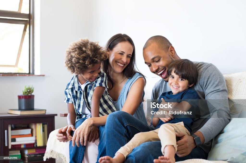 Family having fun at home Happy multiethnic family sitting on sofa laughing together. Cheerful parents playing with their sons at home. Black father tickles his little boy while the mother and the brother smile. Family Stock Photo