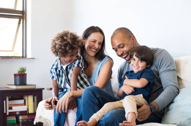 Family having fun at home Happy multiethnic family sitting on sofa laughing together. Cheerful parents playing with their sons at home. Black father tickles his little boy while the mother and the brother smile. multiracial group stock pictures, royalty-free photos & images