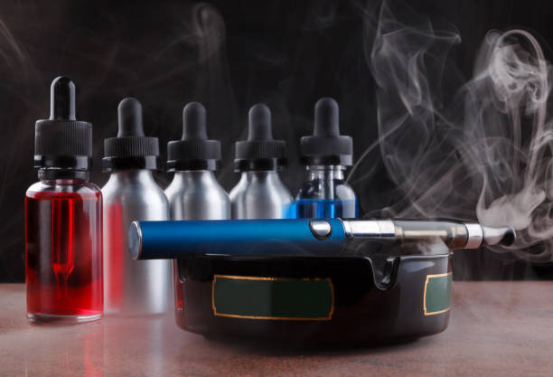 istock Electronic cigarette on the ashtray and vape liquids within vapor on black background 691534064