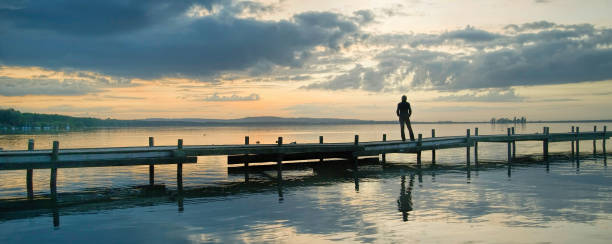 silhouette di donna in piedi sul molo al lago a guardare scenografie idilliache con maestoso paesaggio nuvoloso - steinhuder meer foto e immagini stock