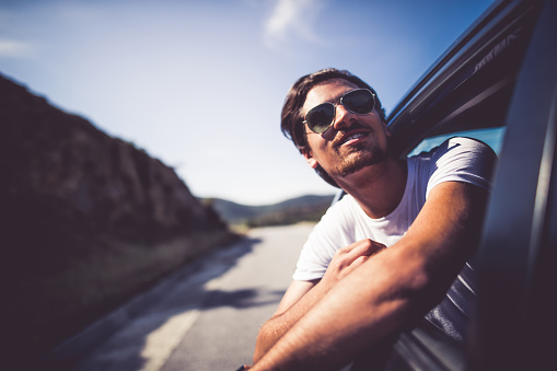 Young hipster man leaning on the car window while driving in the car on the roadtrip and having fun