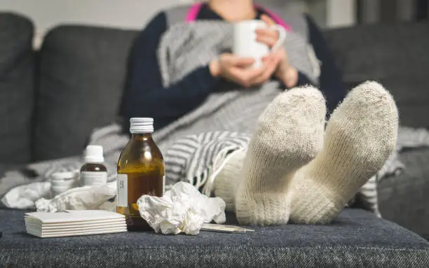 Photo of Cold medicine and sick woman drinking hot beverage to get well from flu, fever and virus. Dirty paper towels and tissues on table. Ill person wearing warm woolen stocking socks in winter.