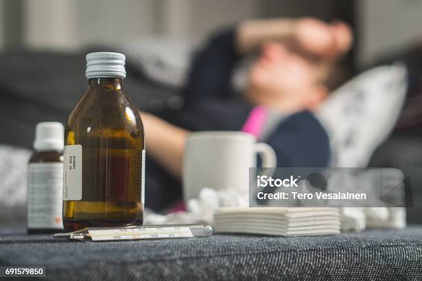 Sick Woman Holding Head And Forehead With Hand And Arm Medicine Thermometer Hot Beverage And Dirty Paper Towels In Front Person Having Flu Fever And Headache On Sofa Couch Caught Cold In Winter Stock Photo - Download Image Now