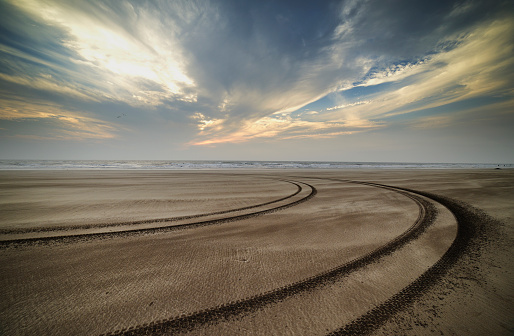 Sandy beach with sea in background at sunset and dramatic cloudy sky with tire tracks going away.