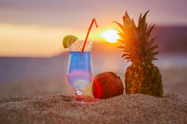 Glass of blue Hawaiian Cocktail with coconut and pineapple on tropical summer scenery, sunset on the beach. Straw in glass.
