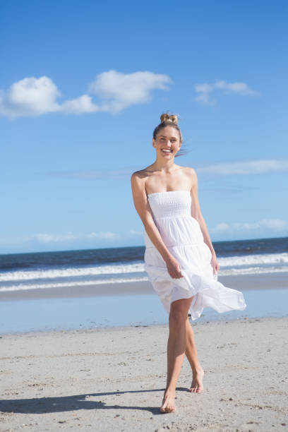 Blonde in white dress walking on the beach smiling at camera on a bright day