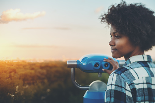 Tilt-shift portrait of meditative young black female with afro curly hair standing in high observation point and holding blue binocular with copy space zone for text, logo or your advertising, sunset