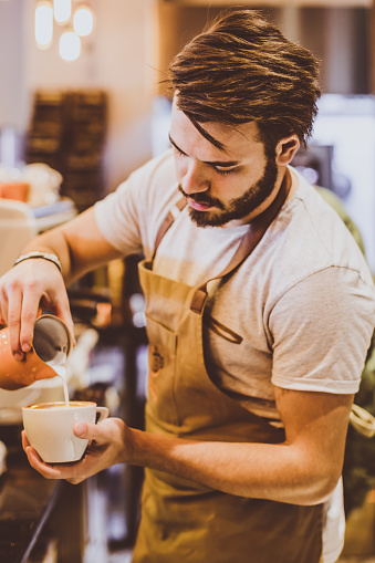 Portrait of young handsome male barista making a cup of coffee and pouring latte art with milk inside of his cafe shop XXXL Size