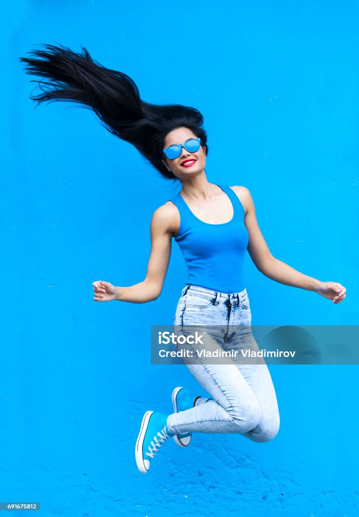 Jumping young woman Jumping young woman in blue in front of blue wall background Blue Background Stock Photo