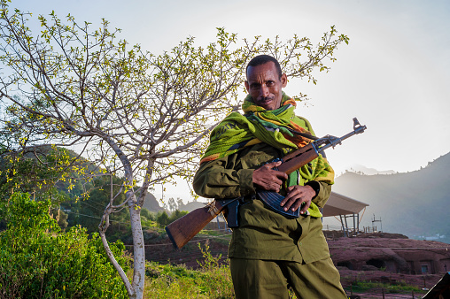 Man guarding with AK47 in full view well lit with green and blue and white sunrise sky behind Lalibela Ethiopia Horn of Africa