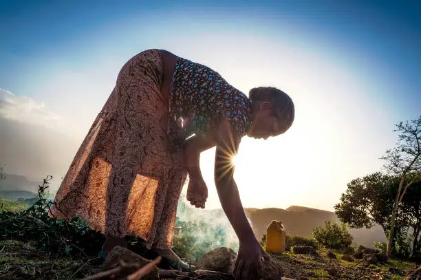 Woman bending over with sun behind her to prepare to perform a Traditional Coffee Ceremony on a hillside outside Lalibela Ethiopia Horn of Africa