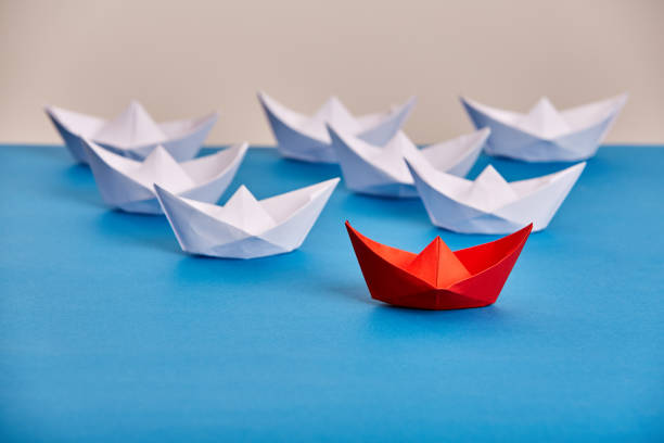 istock Bright red paper ship leading white ones based on blue against light background. 691754140