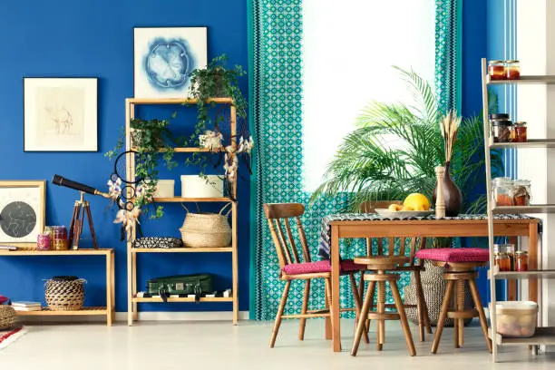 Postcolonial room with wooden bookcase, dining table, chairs, pattern curtain