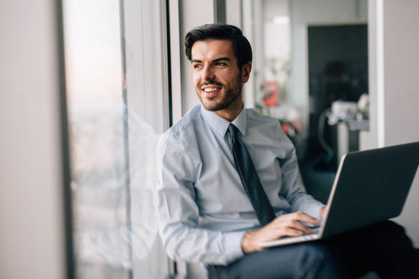 Buisnessman Using Laptop Businessman using a laptop at the window in his office. three quarter length stock pictures, royalty-free photos & images