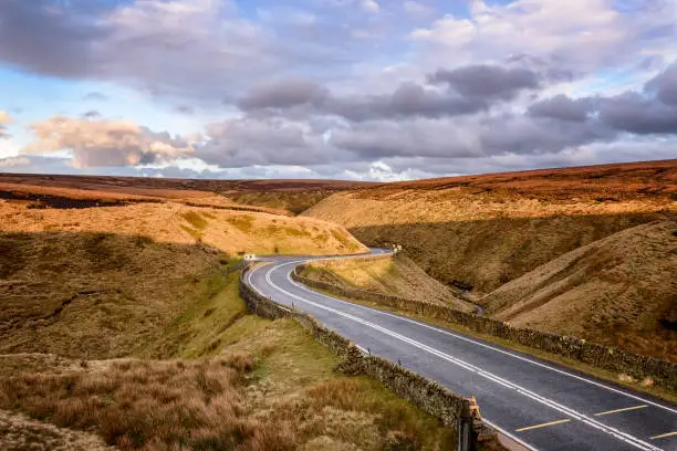 A tarmac road winding through moorland on a cloudy day in Peak District, England.