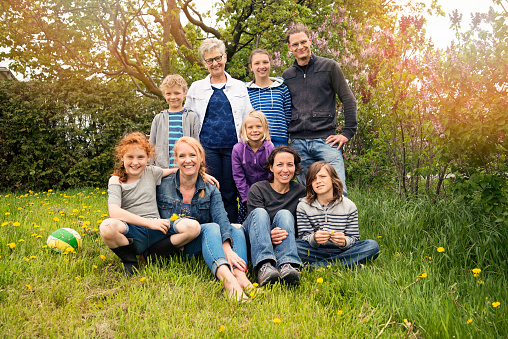 A grandmother, her two mature children, a daughter in law and five grandchildren casually posing outdoors sitting on grass in spring nature. Dandelions and lilacs around. Horizontal full length outdoors shot with copy space. This was taken in Quebec, Canada.