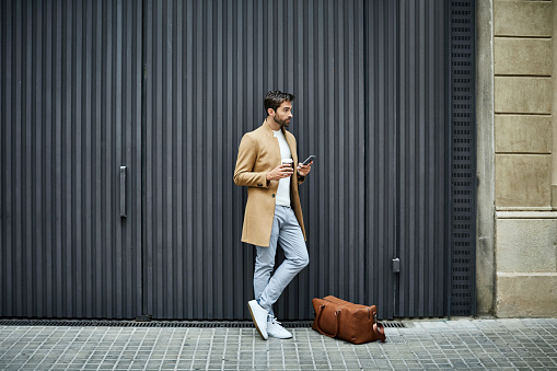 Full length of businessman holding disposable cup and mobile phone. Executive is looking away while standing by bag. He is on sidewalk against building in city.