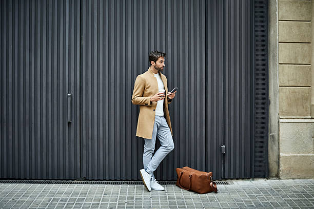 Businessman with phone and cup looking away Full length of businessman holding disposable cup and mobile phone. Executive is looking away while standing by bag. He is on sidewalk against building in city. smart casual stock pictures, royalty-free photos & images