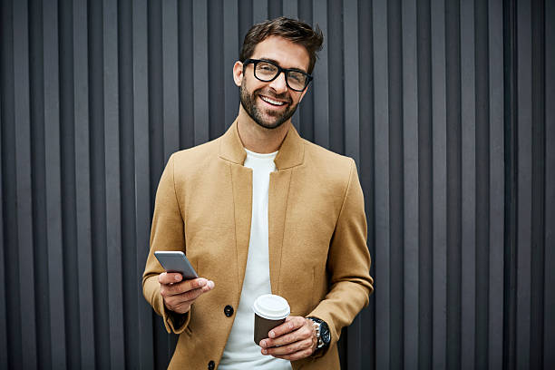 Happy businessman with smart phone and cup in city Happy businessman with disposable cup and smart phone. Portrait of professional wearing smart casuals. He is standing against wall. businessman happiness outdoors cheerful stock pictures, royalty-free photos & images
