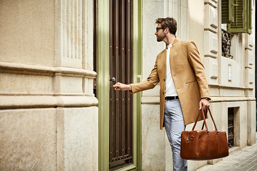 Businessman locking door while standing on sidewalk. Professional is wearing smart casuals. He is carrying brown bag.