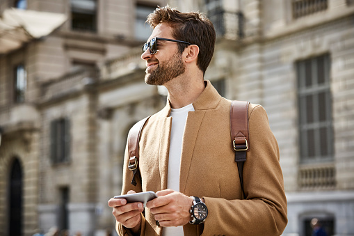 Smiling businessman looking away while holding smart phone. Male professional is wearing smart casuals and sunglasses. He is standing in city.