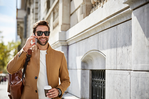 Smiling businessman talking on phone while holding disposable cup. Professional with bag walking by building. He is wearing sunglasses and smart casuals.