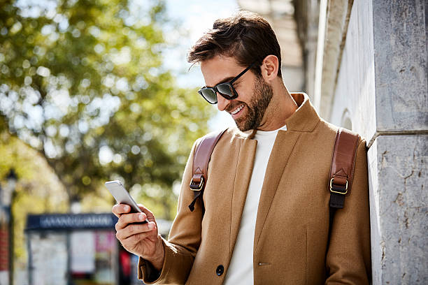 He is carrying bag in city. Smiling businessman using smart phone while leaning on wall. Male executive is wearing smart casuals on sunny day businessman happiness outdoors cheerful stock pictures, royalty-free photos & images