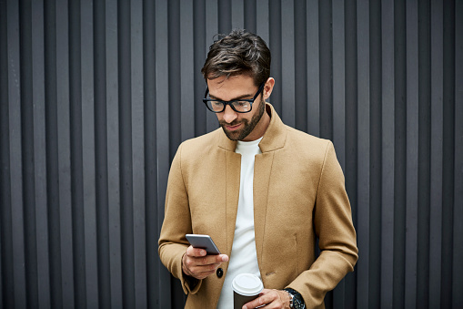 Businessman texting on smart phone and holding disposable cup. Confident executive wearing smart casuals and eyeglasses. He is standing against building.