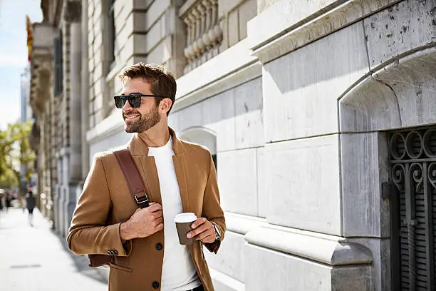 Smiling businessman looking away while holding disposable cup and bag. Professional is walking on sidewalk by building during sunny day. He is wearing smart casuals and sunglasses in city.