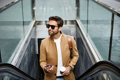 Smiling businessman holding smart phone and disposable cup. Male professional is standing on escalator. He is wearing sunglasses in city.