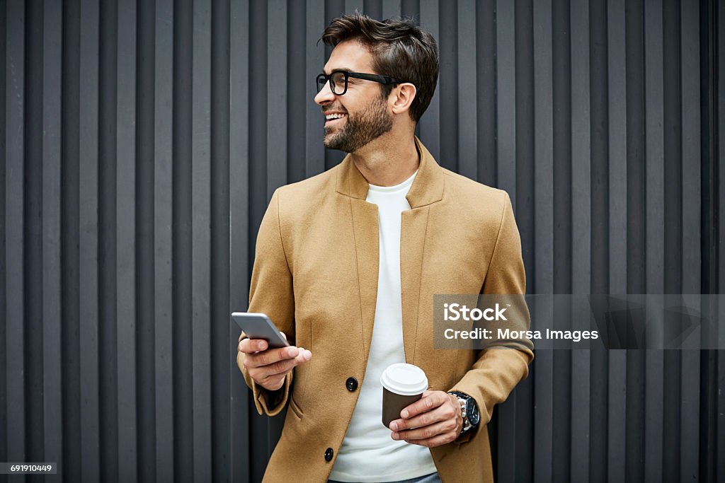 Smiling businessman with smart phone and cup Smiling businessman with smart phone and disposable cup. Handsome executive looking away while standing against wall. He is wearing smart casuals. Men Stock Photo