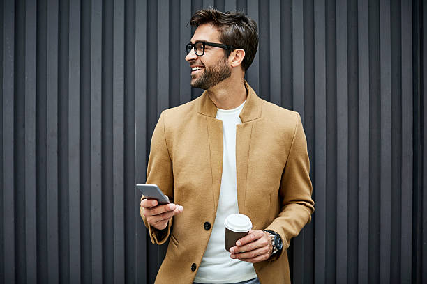 Smiling businessman with smart phone and cup Smiling businessman with smart phone and disposable cup. Handsome executive looking away while standing against wall. He is wearing smart casuals. horn rimmed glasses stock pictures, royalty-free photos & images