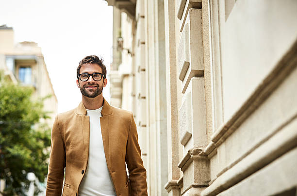 Smiling businessman walking by building in city Smiling businessman walking by building. Confident professional is in smart casuals. He is wearing eyeglasses. businessman happiness outdoors cheerful stock pictures, royalty-free photos & images