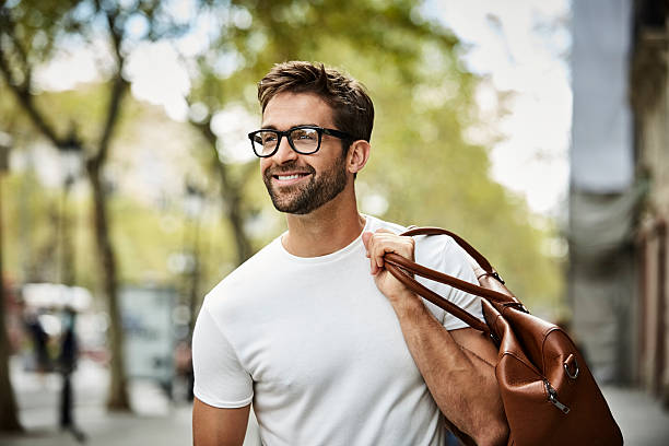 smiling businessman with brown bag walking in city - brille stock-fotos und bilder