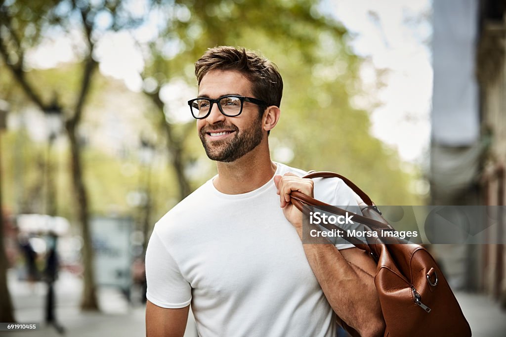 Smiling businessman with brown bag walking in city Smiling handsome businessman with beard walking in city. Executive is looking away while carrying bag. He is wearing eyeglasses. Men Stock Photo