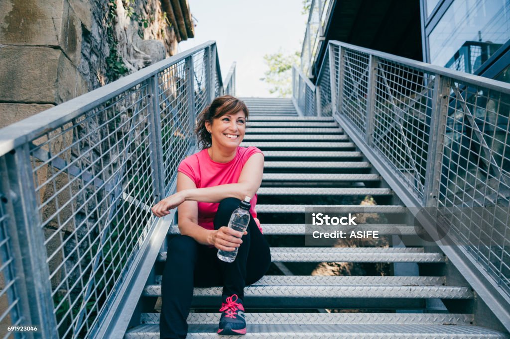 Laugh is the best sport Happy and beautiful middle aged woman sitting on metallic stairs relaxing before running outdoors holding a water bottle Mature Women Stock Photo