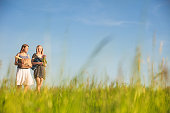 Female friends talking while walking on grassy field against blue sky