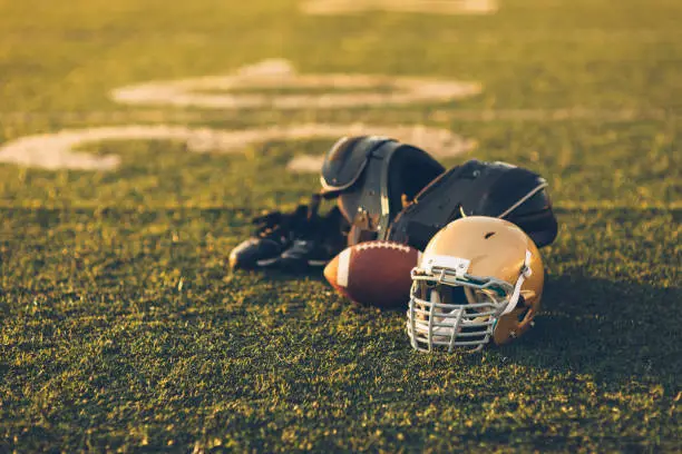 A Gold American Football helmet sits with a football on a football playing field. The light is from the sun which is about to set, shallow depth of field. Copy space included. Sport background image.