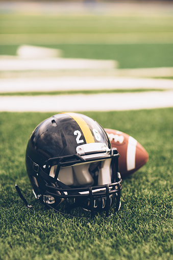 An American Football helmet sits with a football on a football playing field. The light is from the sun which is about to set, shallow depth of field. Copy space included. Sport background image.