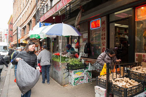 This is a color photograph of an Asian woman carrying a full bag past a Chinatown market in New York City.