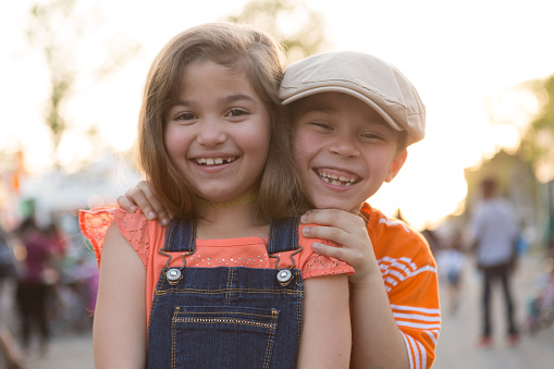 A little girl and boy smile and pose for the camera.  The boy stands behind the girl and peeks out over her shoulder.  Both children are hispanic.  The girl has light brown hair and wears overalls.  She is smiling and missing a tooth.  The boy is smiling and also missing some teeth, he wears a hat.  Shot at dusk, backlit.