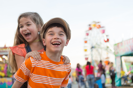 A little girl and boy at a carnival.  The little girl is riding on the boy's back.  They are laughing and shouting.  Both children are hispanic.  The girl has light brown hair and wears overalls.  She is smiling and missing a tooth.  The boy is smiling and also missing some teeth, he wears a hat.