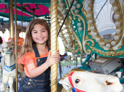 A little girl rides a carousel.  She is smiling and looking at the camera while she rides a wooden horse, she is holding onto the ride.  She is hispanic, has brown eyes, brown hair, and is wearing a pair of overalls with an orange shirt.