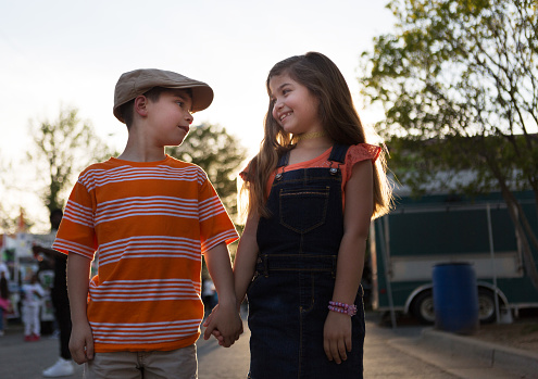A little boy and girl hold hands and look at each other, smiling.  Both children are hispanic.  The girl has long light brown hair and is wearing overalls and an orange shirt.  The boy is wearing an orange and white striped shirt and a hat.  They are outdoors and backlit.