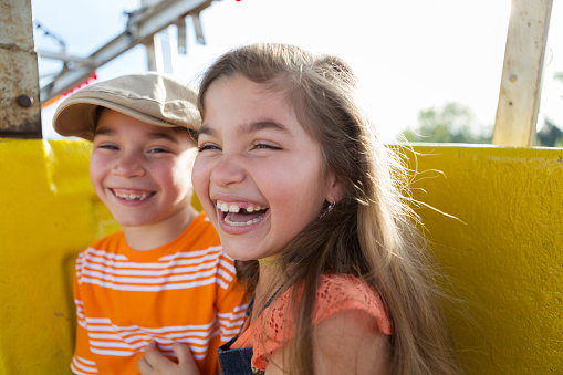 A little girl and boy riding in a ferris wheel together.  The girl is closer to the viewer.  Both are smiling at the camera.  She has long brown hair, brown eyes, olive skin, and is missing a tooth.  Both children are hispanic and wearing orange shirts.  The girl is laughing.